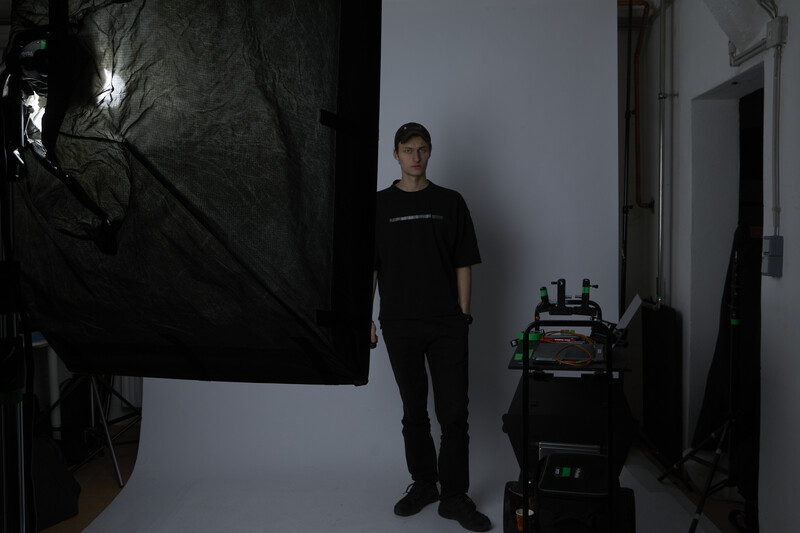 Portrait subject standing in studio with professional lighting setup and equipment visible.