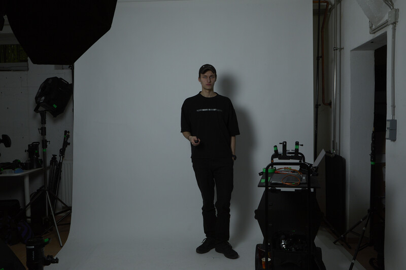 Man in black clothing standing against white wall in photography studio with professional lighting equipment visible.