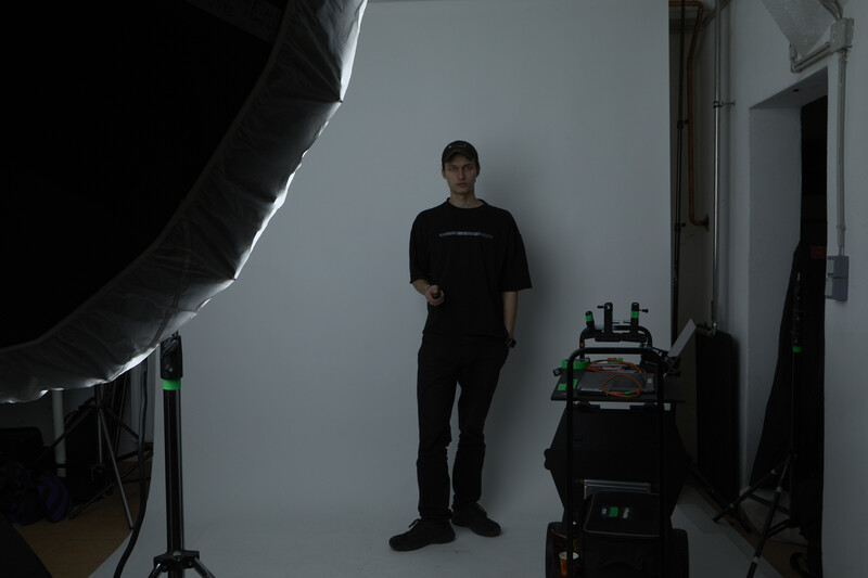 Man in black clothing standing against white backdrop during studio photo session with professional lighting equipment visible.