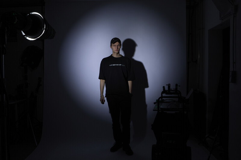 Man in black shirt standing in dramatic studio lighting with circular light source and shadow cast on backdrop.