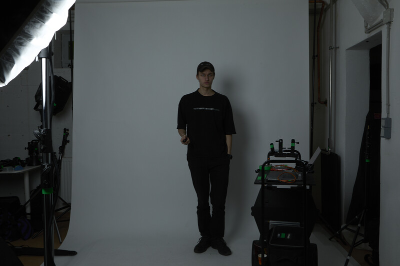 Person standing against white backdrop in professional studio with lighting equipment and cases visible.