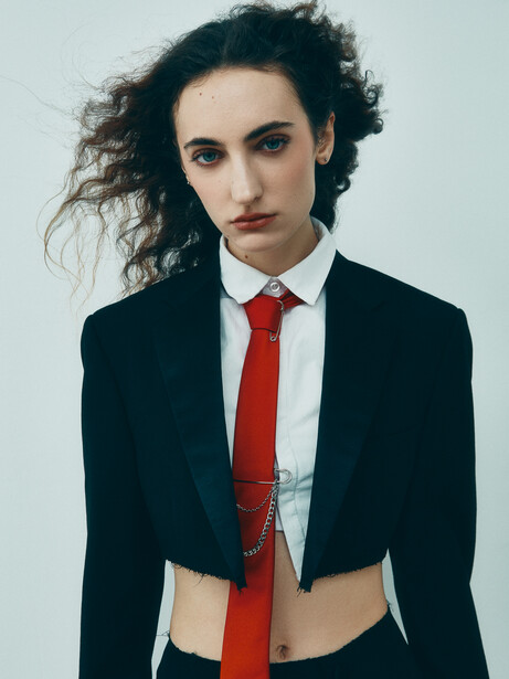 Portrait of a person in cropped black blazer and red tie against neutral background.