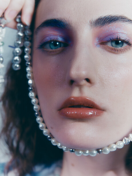 Close-up portrait with purple and blue eyeshadow, beaded necklace, and direct gaze at camera.