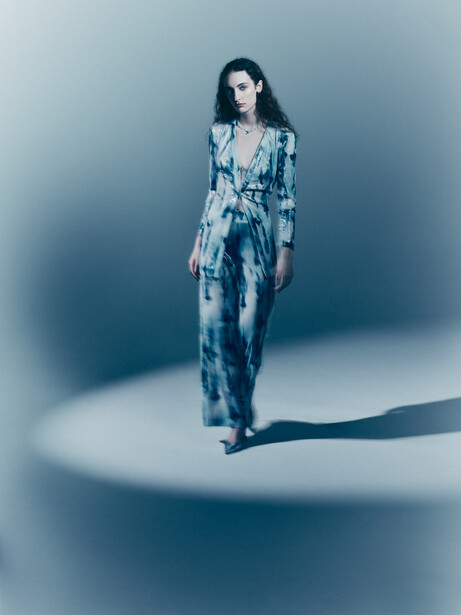 Woman in patterned blue outfit standing in minimalist studio with dramatic side lighting and shadow.