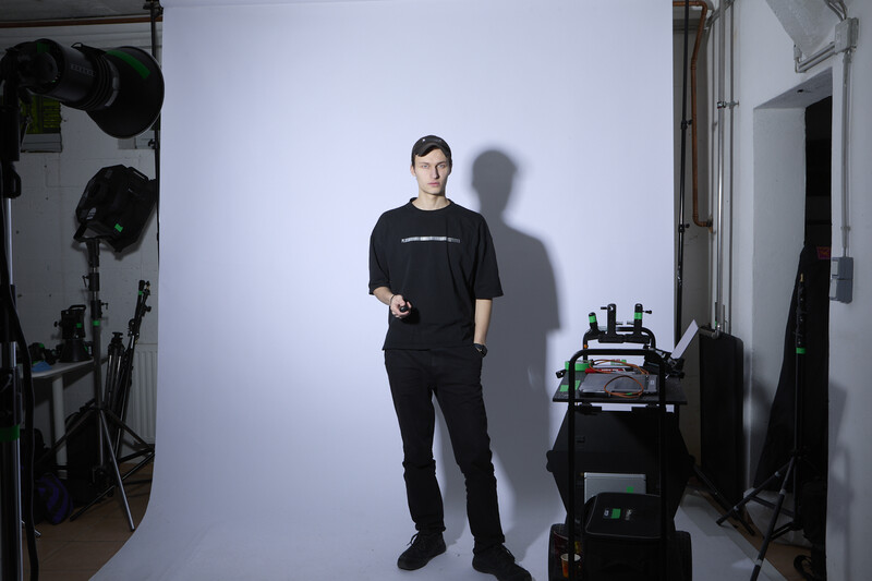 Man in black shirt and pants standing in professional studio with white cyclorama backdrop and lighting equipment.