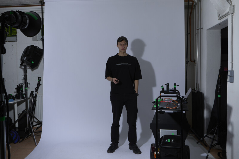 Man in black clothing standing in a studio with professional lighting equipment and camera gear visible.