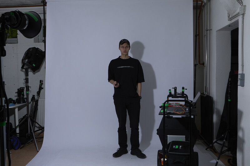 Man in black clothing standing in a professional studio setup with white backdrop and studio lighting equipment.