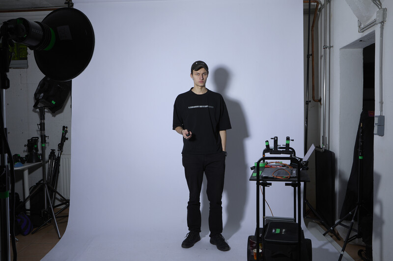 Man in black clothing standing in a studio with professional lighting equipment and a wheeled cart visible.