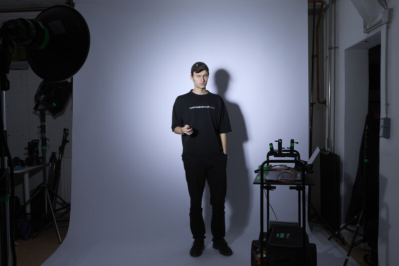 Person in black clothing standing against a bright backdrop in a professional studio setup with lighting equipment visible.
