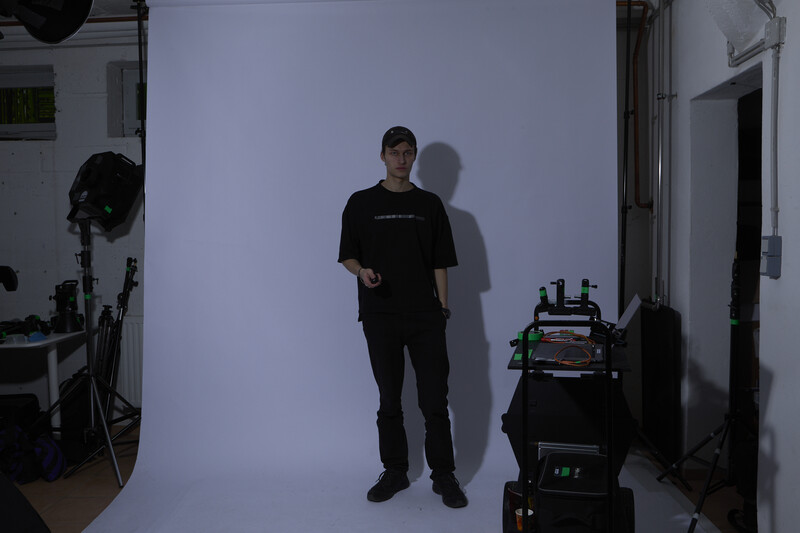 Man in black clothing standing against white backdrop in studio with professional lighting equipment visible.