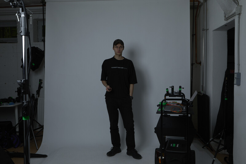 Man in black clothing standing against white studio wall with professional lighting equipment visible.