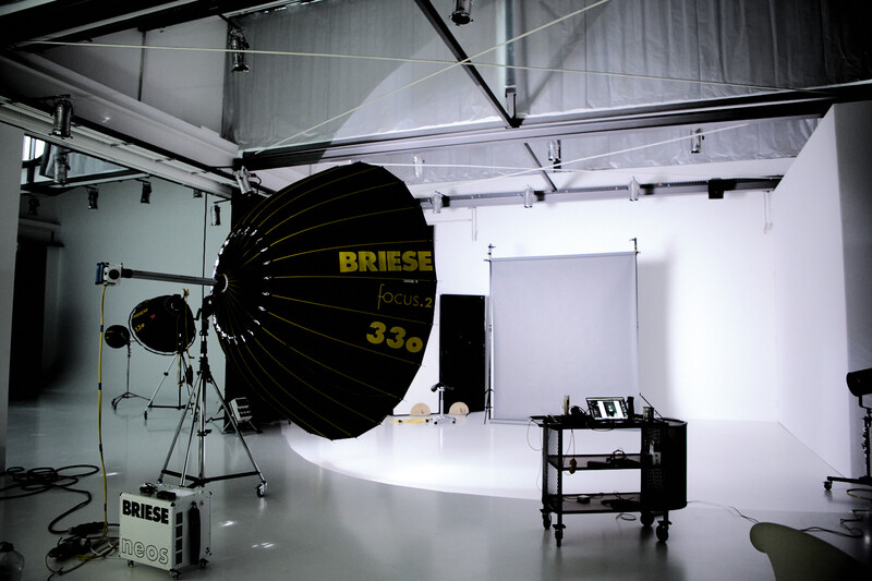 Professional photography studio with a large parabolic softbox and key light positioned above a white seamless backdrop.