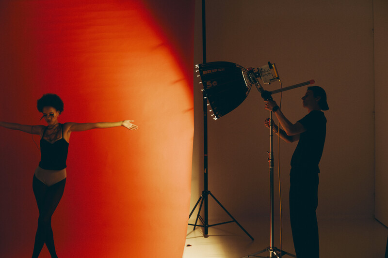 Two people posing during a studio photoshoot with professional lighting equipment and a warm red-orange gradient backdrop.