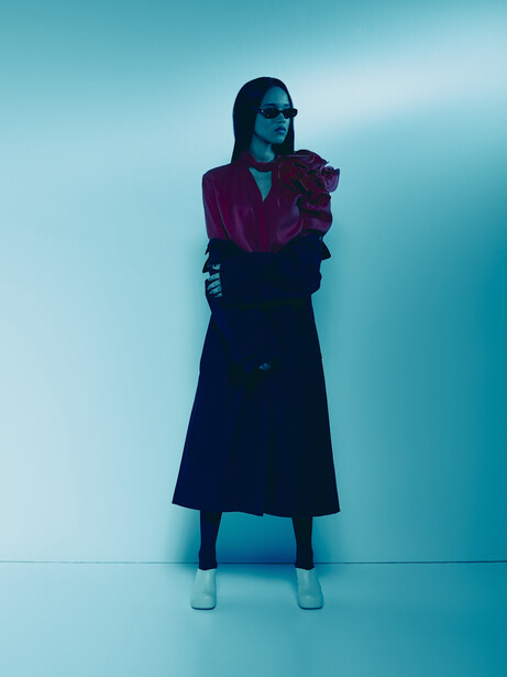 Woman in burgundy jacket and navy skirt standing against blue gradient backdrop.