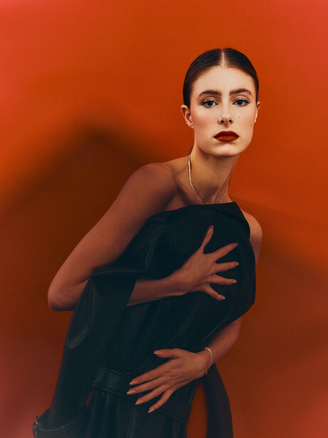 Woman in dark clothing posed against vibrant orange background with dramatic side lighting and shadow.