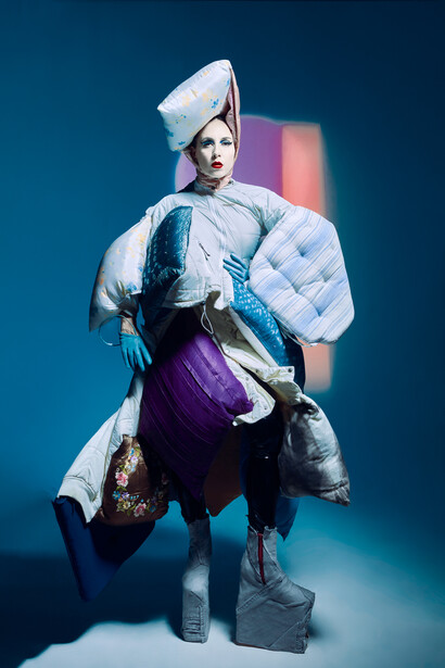 Person in avant-garde outfit with white turban and quilted jacket against blue studio background.