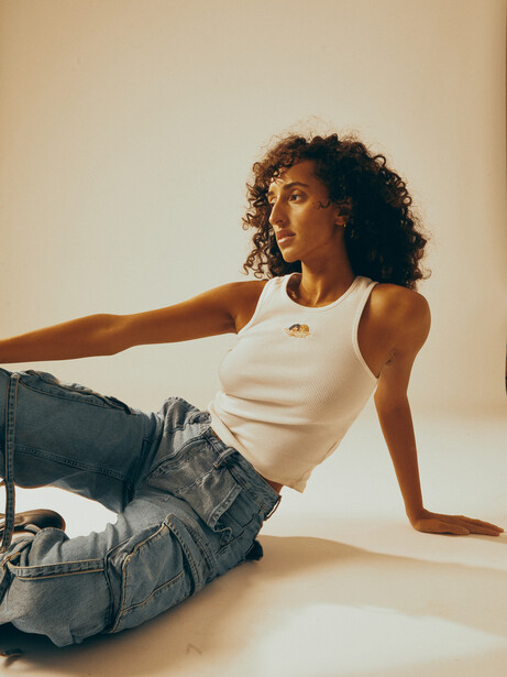 Woman in white tank top and jeans reclining on neutral background with extended arm.