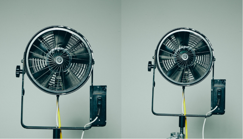 Two industrial floor fans mounted on stands with yellow power cords against a plain white wall.