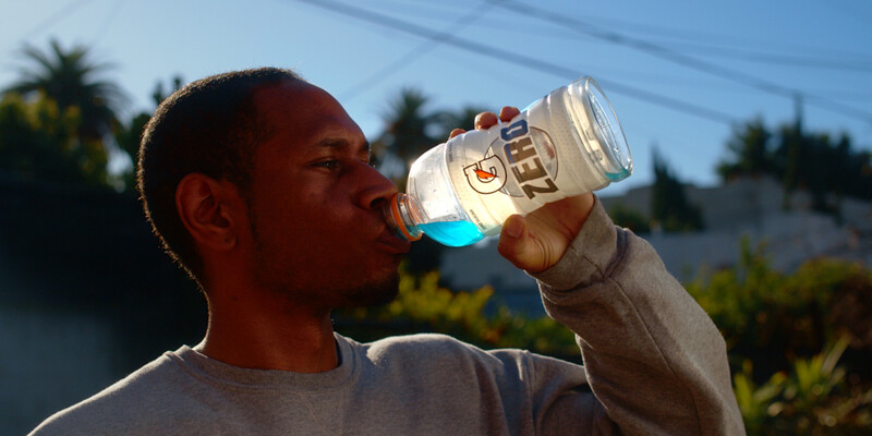 Man in gray shirt drinking from a clear bottle with blue liquid against a bright outdoor background.