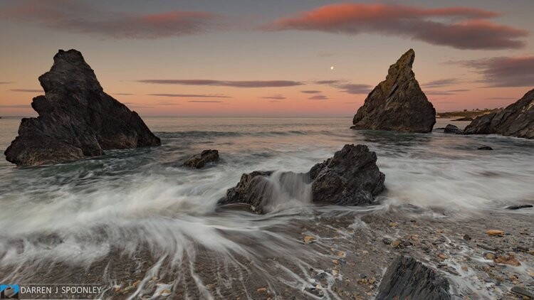 Rocky sea stacks rise from foaming waves during twilight, with a crescent moon visible in the dusky sky.