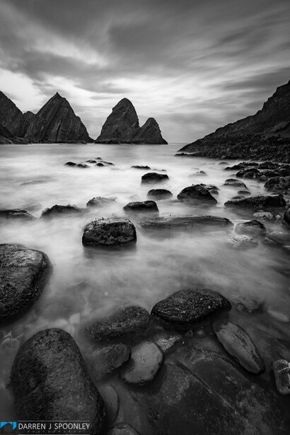 Rocky coastline with sea stacks and boulders in misty water under dramatic cloudy sky.