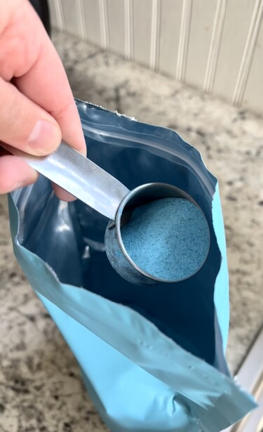Hand holding a spoon with blue powder over an open bag of blue substance.
