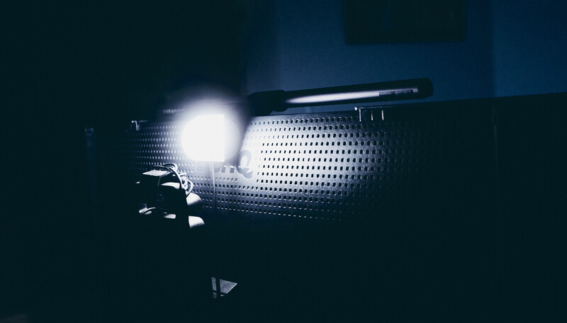 Close-up of a perforated metal speaker or grille with a bright light source illuminating its surface against a dark background.