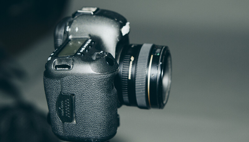 Professional DSLR camera with attached lens, photographed against a neutral gray background with dramatic side lighting.