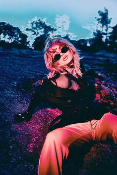 Person wearing pink sunglasses and dark clothing sits in a moody landscape with neon pink and blue lighting.