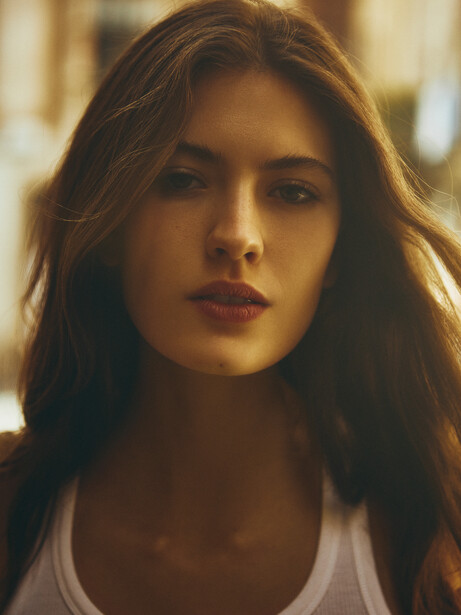Woman with long dark hair wearing a white tank top, photographed indoors with warm golden lighting.