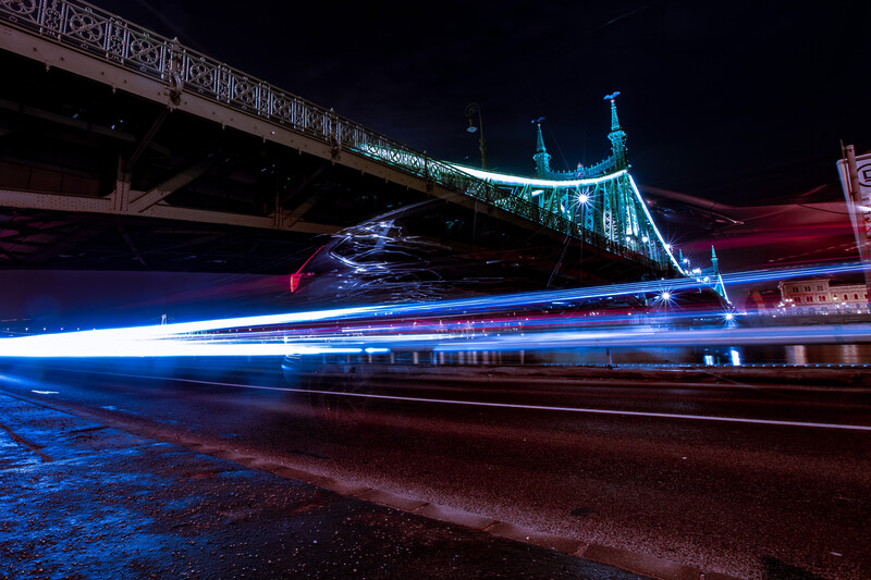 Long exposure photograph of a bridge illuminated with blue and cyan lights, with a blue light trail from traffic streaking across the frame.