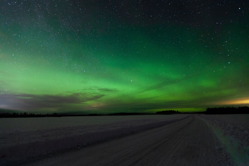 Northern lights glow green across night sky above a frozen lake and distant forest.