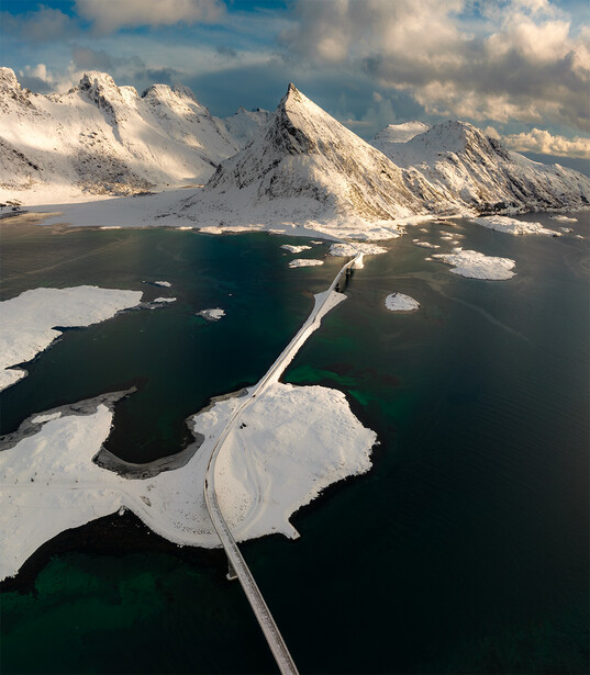 Aerial view of snow-covered mountains and a causeway crossing a dark water channel with ice floes.