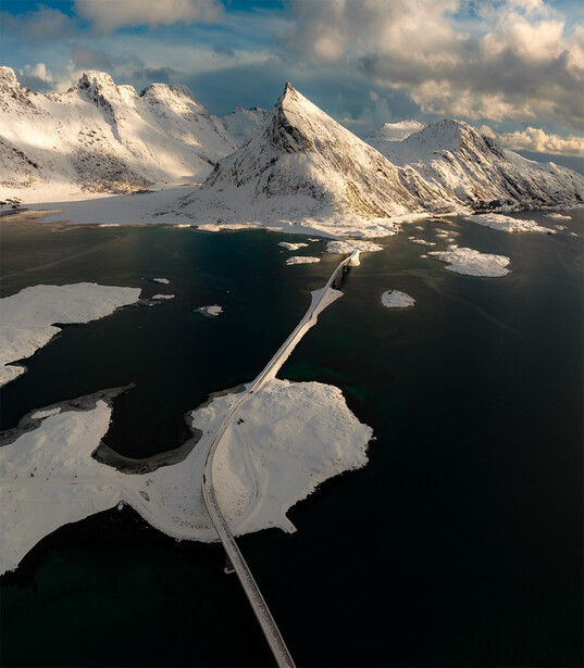 Aerial view of snow-covered mountains and dark water with ice formations and a bridge connecting islands.