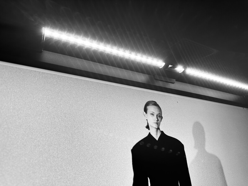 Person in dark clothing against white wall with strong side lighting and visible shadow.