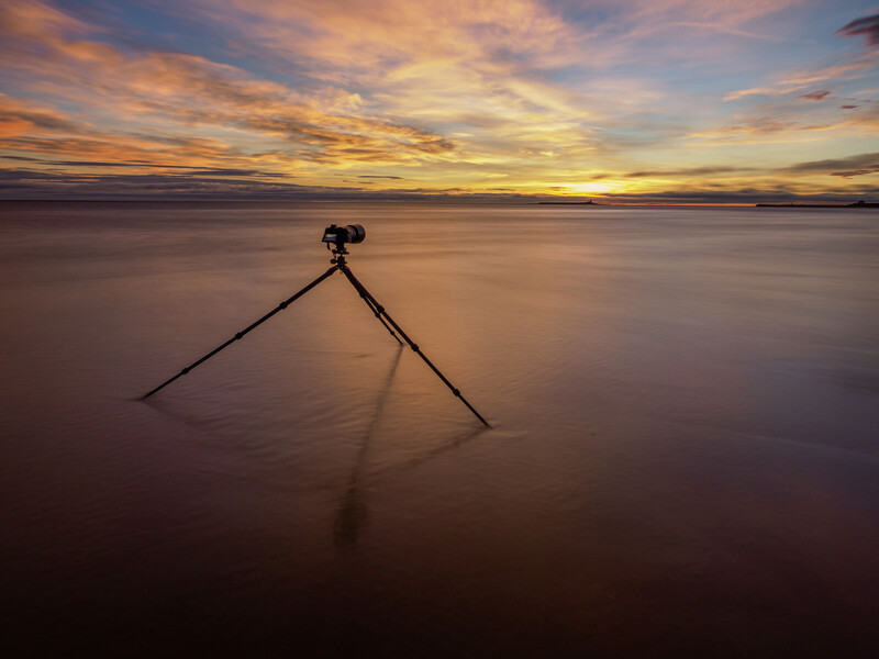 Tripod standing alone in shallow water at sunset with golden sky reflected below.