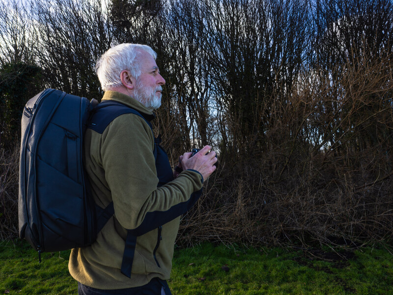 Older man with white hair jogging outdoors wearing a blue and navy backpack and olive jacket.