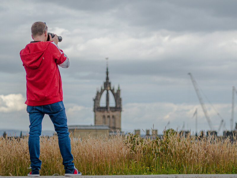 Child in red jacket photographing a distant stone tower across a harvested grain field.