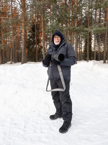 Person in winter clothing holding a camera in a snowy forest clearing.