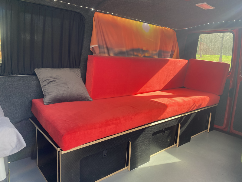 Red fabric-covered daybed or seating platform in a studio or van interior with dark curtains and overhead lighting.