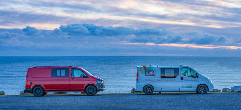 Two camper vans parked side-by-side on a coastal road at dusk, with calm ocean and dramatic sky beyond.