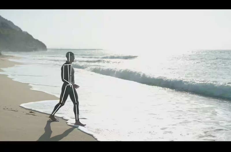Surfer in wetsuit standing on beach holding surfboard, facing ocean with waves rolling in.
