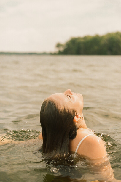 Woman in water looking upward, shot from behind with soft natural lighting and blurred shoreline background.