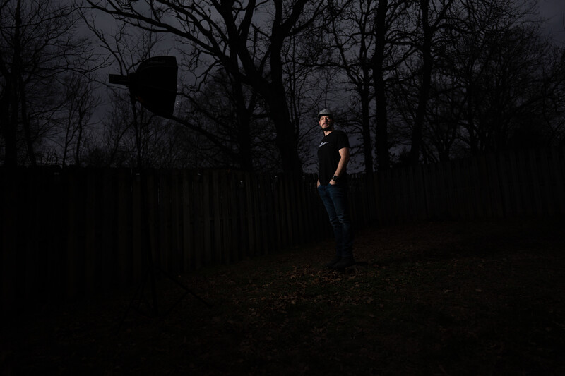 Person standing in dark woods at night, illuminated by single light source against bare trees and wooden fence.