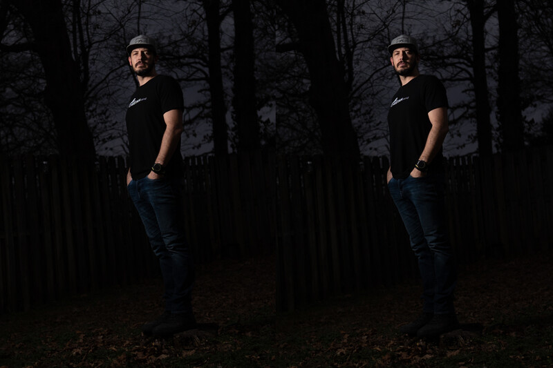 Man in black t-shirt and cap standing in dark forest setting at dusk.