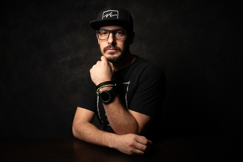 Man wearing glasses and black cap rests chin on fist in contemplative pose against dark background.