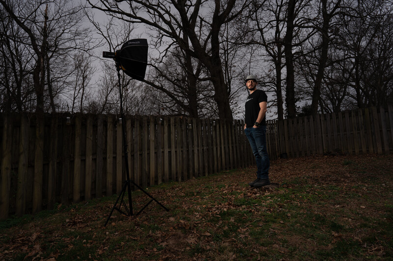 Photographer standing in a backyard with bare trees, adjusting a large reflector mounted on a light stand.