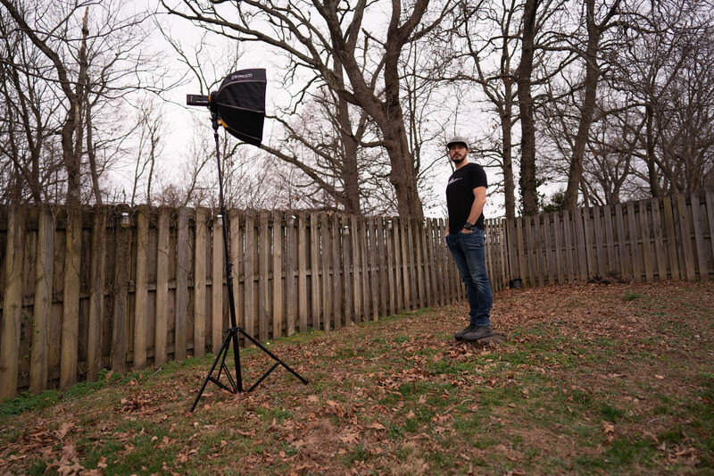 Photographer practicing outdoor lighting setup with reflector on tripod in wooded area.