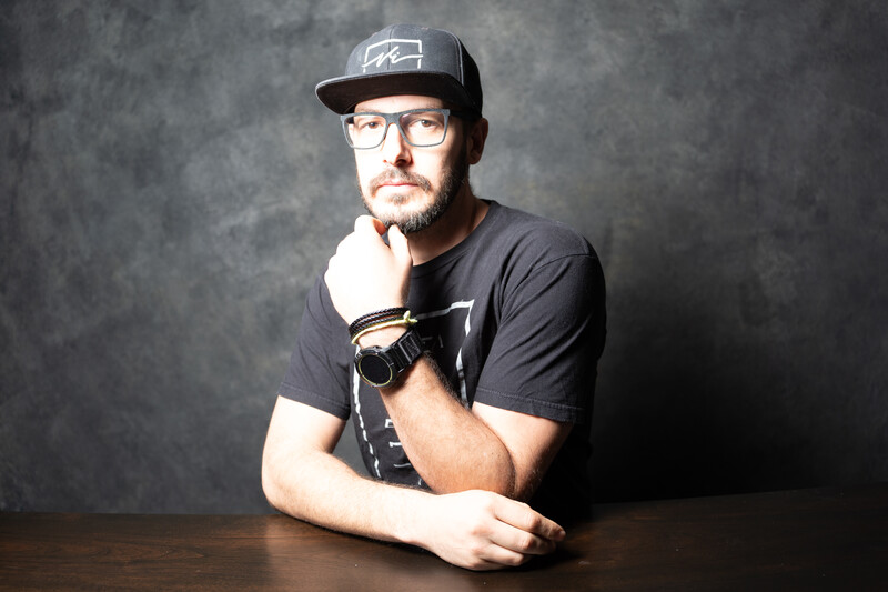 Man wearing glasses and cap with hand raised to chin in thoughtful pose against textured gray background.
