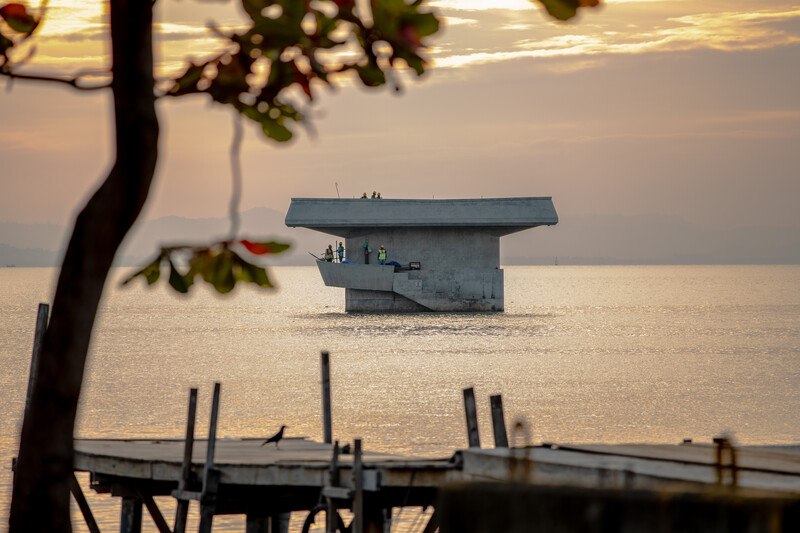 Weathered boat house on calm water at sunset, framed by tree branch in foreground.